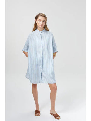 A person wearing a loose, knee-length LANHTROPY - LIGHTHOUSE LINEN SHIRT DRESS made from premium linen in light blue and white stripes stands against a plain white background. The shirt dress from LANHTROPY features buttons down the front. The person has long hair and is wearing brown sandals.