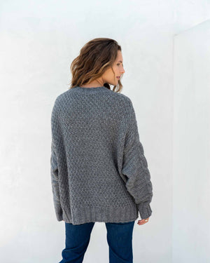 The image displays a detailed view of a person dressed in the MERSEA - TOPANGA CABLE SWEATER by MER SEA, showcasing its chunky knit cream-colored texture with large buttons on the sides, paired with blue jeans. The emphasis is on the sweater's intricate cable design and fine details, with the person's hand partially visible at their side.