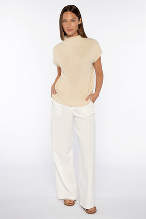 A person in the KINROSS Cap Sleeve Mock Neck Top in a versatile beige cotton blend and white wide-leg pants stands against a plain white background, hands in pockets, wearing brown sandals.