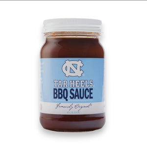 A jar of Cackalacky - Carolina Tar Heels BBQ Sauce is placed on a wooden surface, alongside a light blue football helmet and football. The display is artistically enhanced with scattered blue and white paper shreds, highlighting the vibrant appeal of this savory tomato-based barbecue sauce from Cackalacky.