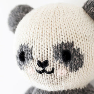 Introducing the CUDDLE + KIND - HANDMADE BABY PANDA from CUDDLE & KIND, a charming, hand-knit plush toy featuring gray ears, arms, and legs, and a white body accented with gray around the eyes and limbs. The panda's smiling face and black eyes add to its adorable appeal. This lovely toy is perfect for baby shower gifts, crafted with love to support fair trade income initiatives.