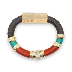 The HOLST & LEE - Luxe Colorblock Bracelet showcases sections of black, teal, and red thread, accentuated by gold-tone rings and a central rhinestone-studded bead. The gold-plated clasp is engraved with "HOLST" and "LEE" labels.