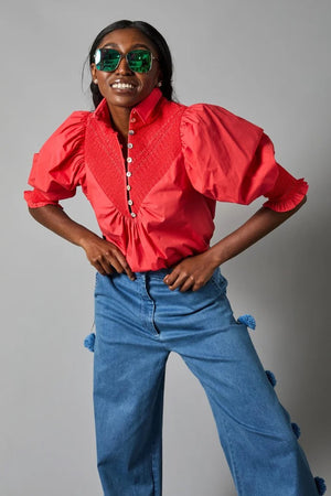 Rear view of a person wearing the Hope for Flowers Smocked Shirt, a bright red blouse with puffed sleeves, paired with blue jeans featuring pom-poms. Made from GOTS Certified organic cotton, this outfit embodies sustainable fashion against a gray background.