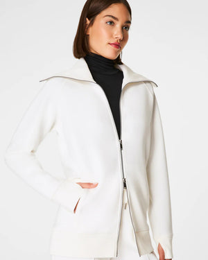 Someone with shoulder-length hair wears a white SPANX AirEssentials Luxe Full Zip Tunic Jacket with matching pants, hands in the tunic's pockets. The plain white backdrop highlights the minimalist loungewear aesthetic.