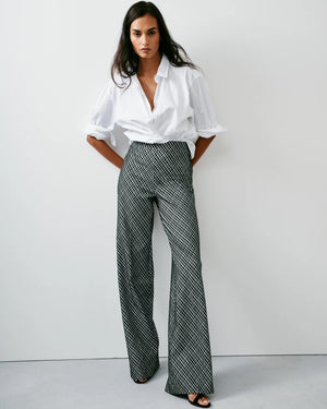 A person is wearing SPANX's Ponte Jacquard Wide Leg Pant, designed with a high-waisted fit and showcasing a black and white grid pattern crafted from premium fabric. They are paired with a black top, standing with their right arm by their side. The focus is on the stylish pants by SPANX.