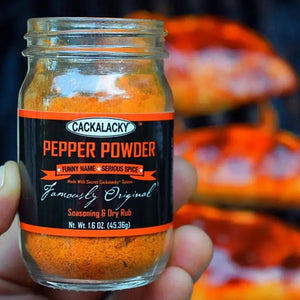 A glass jar of CACKALACKY - PEPPER POWDER RUB, made with fiery chile peppers and secret spices, is shown against a white background. The jar has a black lid and a black label with orange and white text stating "Cackalacky Pepper Powder," "Funny Name - Serious Spice," and "Famously Original." The jar contains 2.3 oz (65.2g).