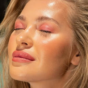 A close-up of a person with curly dark hair, a serious expression, and warm makeup featuring the universally flattering, weightless JASON WU - HOT FLUFF LIP/EYE/CHEEK by JASON WU BEAUTY on their face. They are looking directly at the camera with a neutral background.