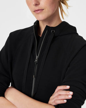 A person with blonde hair is wearing the SPANX AirEssentials Full Zip Hoodie made from ultra-soft fabric, with their hands in the pockets and a black skirt. They are standing against a plain, light-colored background and smiling softly.