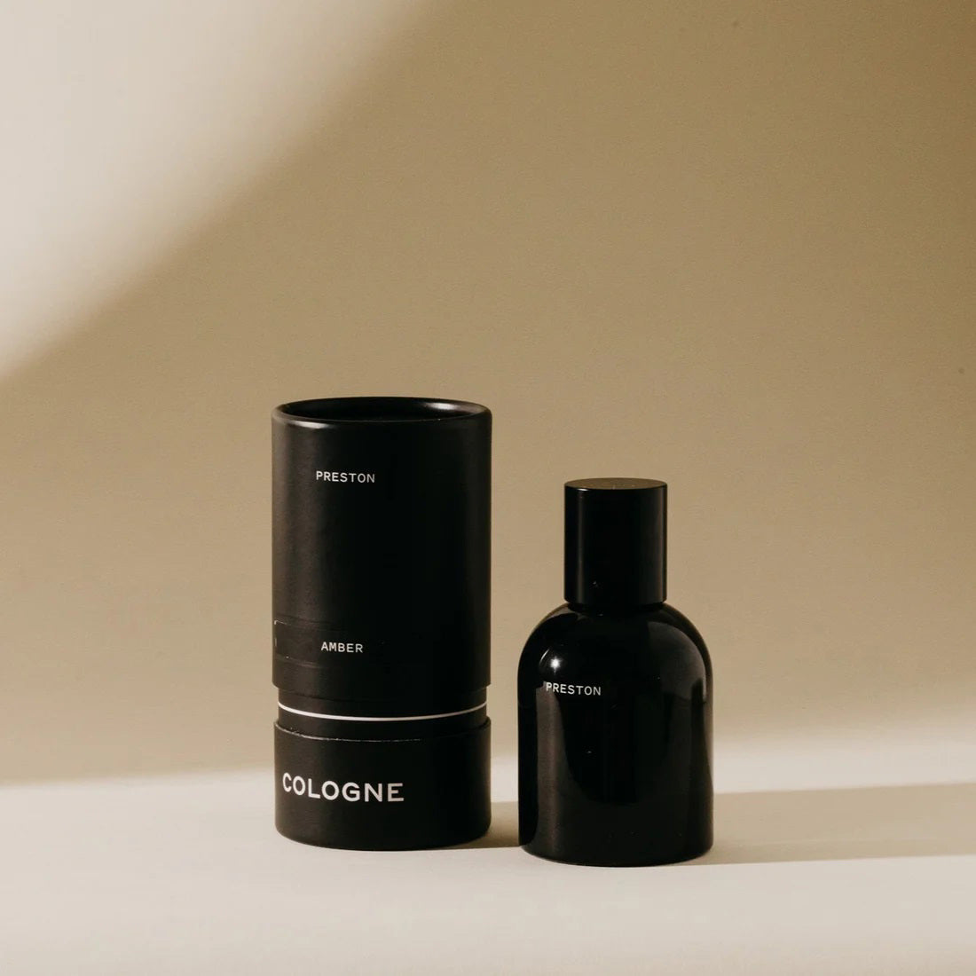 A sleek black bottle of PRESTON - COLOGNE 1.7OZ stands next to its matching cylindrical case. Both items are set against a neutral beige background, creating a minimalist and sophisticated look. The black packaging, made from biodegradable materials, features simple white text labeling the product with the brand name PRESTON.