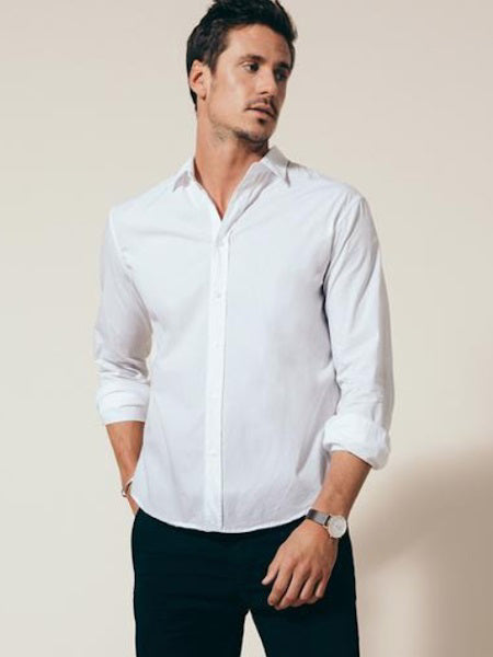 A man in the FRANK &amp; EILEEN Finbar Men&#39;s Button Up Shirt in classic white poplin stands with rolled sleeves and hands in pockets, looking to the side. He has short dark hair, wears a watch, and poses against a light background.