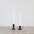 Two lit taper candles in rustic, dark Creative Co-Op hand-forged cast iron holders on a wooden surface, with the left holder taller than the right. The background is a plain light-colored wall.