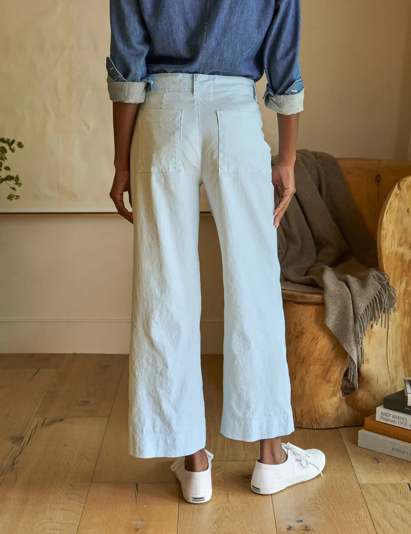 A person in a denim shirt and FRANK AND EILEEN - WEXFORD WIDE LEG LINEN PANT with white sneakers stands with their back to the camera. Nearby, a wooden chair holds a blanket, and books are stacked on the floor.