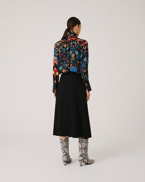 A person with dark hair is standing against a plain background. They are wearing a long-sleeve blouse with a colorful, intricate pattern paired with an elegant SFIZIO - BLACK SKIRT WITH LACE INLAY by SFIZIO. Their outfit, finished with knee-high snakeskin boots made in Italy, looks chic as they look directly at the camera.