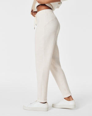 A person stands against a white background, smiling and looking down with one hand touching their hair. They are wearing a matching light beige SPANX AIRESSENTIALS TAPERED PANT and sweatshirt set made from ultra drapey, lightweight spacer fabric, paired with white sneakers.