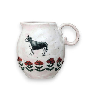 The DOG & FLOWERS HAND PAINTED STONEWARE CREAMER PITCHER by CREATIVE COOP is a white ceramic pitcher adorned with a black dog illustration and a decorative row of red and green floral patterns around the lower portion. It features a curved handle, slightly textured surface, and comes packaged sustainably.