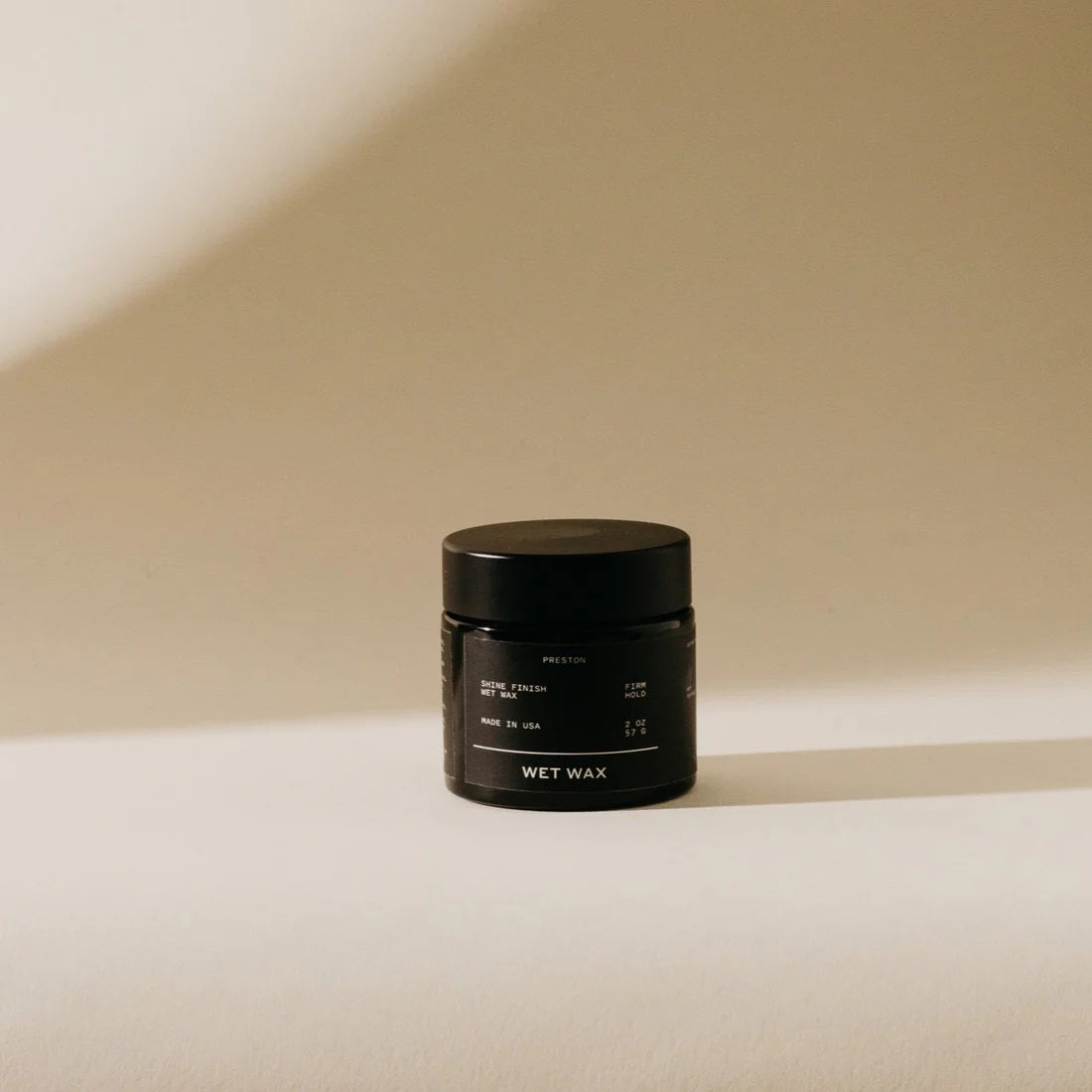 A small, black jar labeled &quot;PRESTON - WET WAX&quot; stands against a plain, beige background. Light and shadow create a soft, minimalist aesthetic around the jar, emphasizing its simple and elegant design. This cruelty-free medium shine product is a firm hold styling aid perfect for sleek finishes.