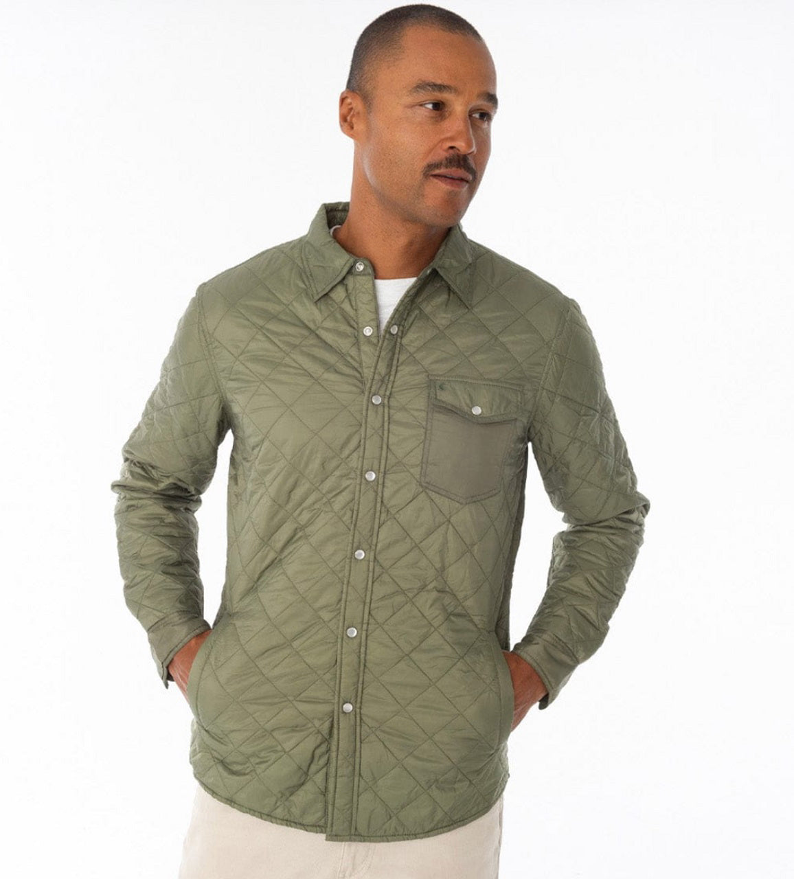 A smiling bearded man stands outdoors in a grassy field, wearing the CRIQUET Quilted Shacket by CRIQUET, paired with jeans and holding a canned drink. Mountains and a wooden fence can be seen in the background.