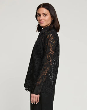 A woman with dark hair stands against a plain background, wearing the HINSON WU - MARGOT LACE SHIRT JACKET, featuring intricate lace detailing and satin trim. She has elegantly paired it with dark pants, her profile turned slightly sideways as she offers a subtle smile.