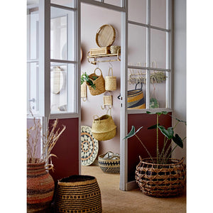 A neatly arranged shelf and stool are adorned with CREATIVE COOP SEAGRASS BASKETS WITH HANDLES, featuring circular handles. These baskets, along with a blanket, a hat, a plant, and decorative pottery against a white paneled wall, create a warm, rustic vibe with natural tones and textures.