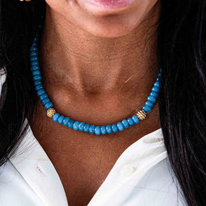 Close-up image of a woman wearing the CAPUCINE DE WULF - BERRY SINGLE STRAND NECKLACE, made of blue beads and pearls with gold spacers. She is wearing a white top. Only the lower part of her face and the upper part of her chest are visible.