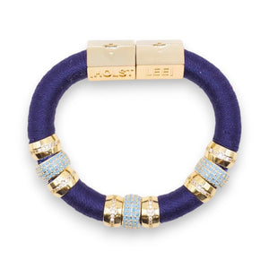 The HOLST & LEE - Luxe Colorblock Bracelet showcases sections of black, teal, and red thread, accentuated by gold-tone rings and a central rhinestone-studded bead. The gold-plated clasp is engraved with "HOLST" and "LEE" labels.