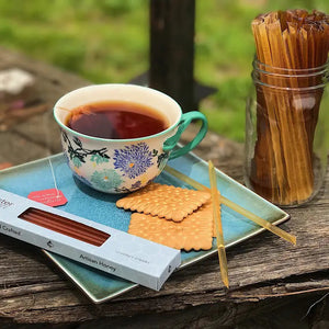 A floral teacup filled with tea sits on a square blue plate with two rectangular biscuits and an open Cloister Honey - Wildflower Honey Straw. A box labeled "Artisan Honey" and a jar containing more Wildflower Honey Straws are beside the plate on a rustic wooden surface with a grassy background.