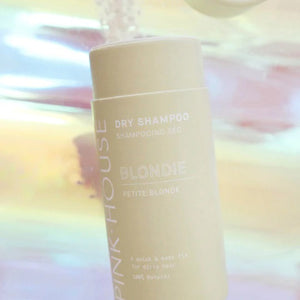 A cylindrical container labeled "PINK HOUSE ORGANICS - DRY SHAMPOO BLONDE" from "PINK HOUSE ORGANICS" is displayed against a colorful, blurred background. Ideal for greasy hair, this non-scented dry shampoo features a light-colored design with minimalist text. The background showcases soft pastel hues.