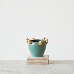 CREATIVE COOP's PLANTER WITH FROGS, a teal ceramic pot with a reactive glaze and beige frog-like figures on the rim, rests on a stack of books atop a marble surface against a light gray background, capturing a subtle farmhouse garden vibe.