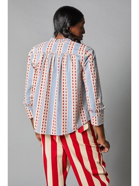 A person with long hair faces away, showcasing the HOPE FOR FLOWERS Frilled Cuff Shirt made from organic cotton, featuring blue and red stripes with dots, paired stylishly with eco-friendly red and white striped pants.