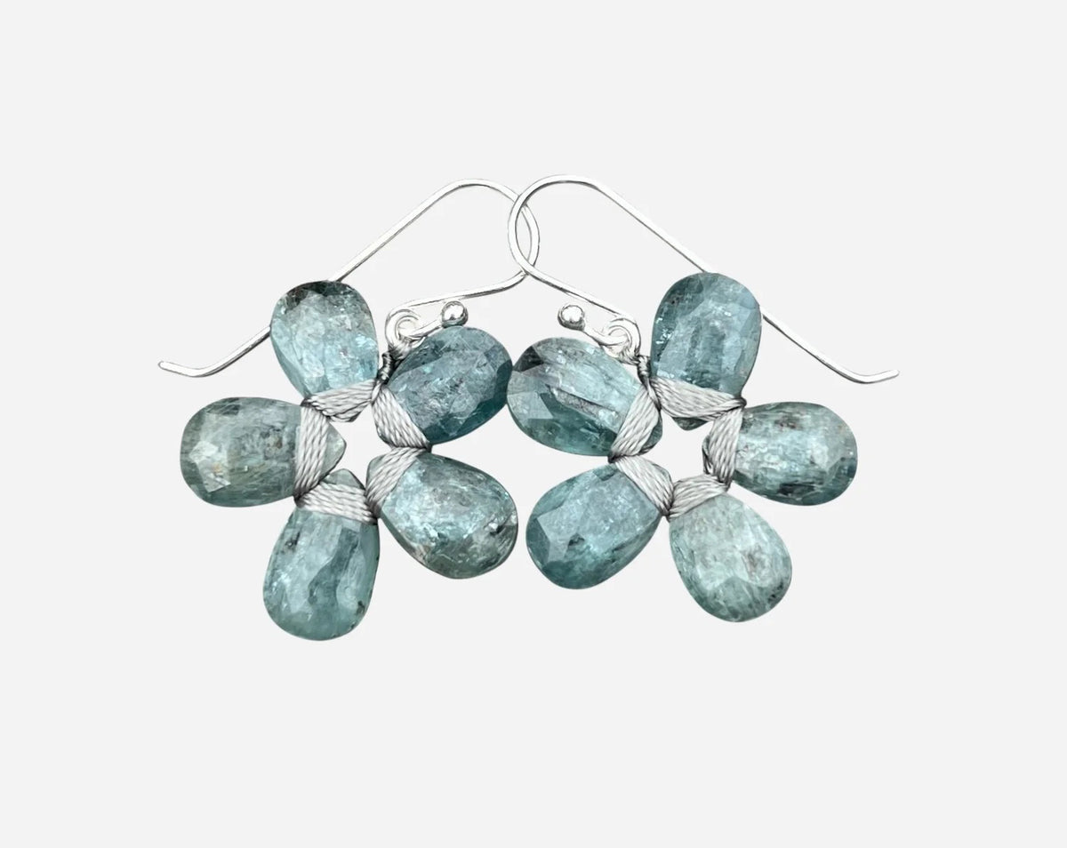 The ANN LIGHTFOOT - BLUE GREEN KYANITE FLOWER EARRINGS by ANN LIGHTFOOT, featuring faceted blue green kyanite clusters, are displayed on a weathered gray wooden surface.