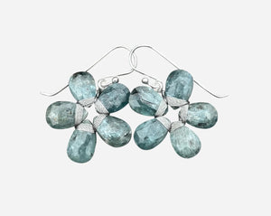 The ANN LIGHTFOOT - BLUE GREEN KYANITE FLOWER EARRINGS by ANN LIGHTFOOT, featuring faceted blue green kyanite clusters, are displayed on a weathered gray wooden surface.