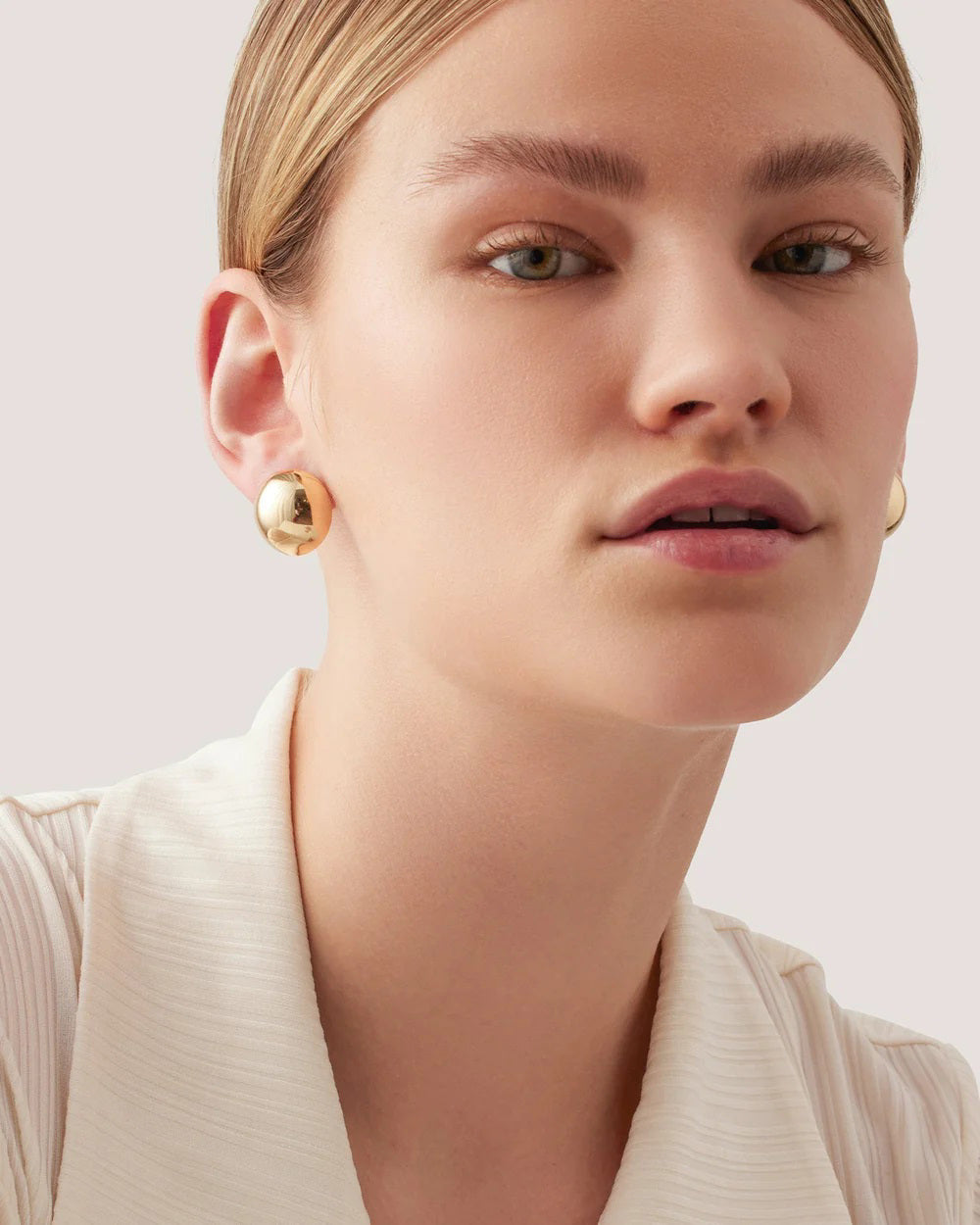 A person with light skin and blond hair is wearing the JENNY BIRD - AURORA EARRINGS, which are round and have a high-polish finish, along with a white collared shirt. They have a neutral expression and are gazing slightly past the camera. The background is plain and light-colored.