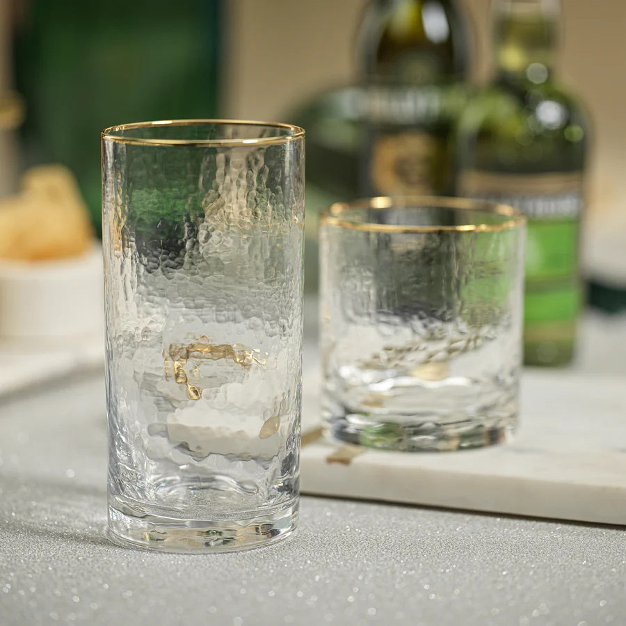 Two textured glass tumblers with gold rims sit on a sparkling surface. The taller, cylindrical tumbler, identified as the NEGRONI HAMMERED HIGHBALL GLASS by ZODAX, has a capacity of 560 ml, while the shorter and wider one holds less. Blurred green bottles and a white bowl in the background enhance the subtle and elegant atmosphere.