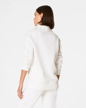 A person with dark hair stands against a plain white background, wearing the super-soft SPANX AirEssentials Luxe Half Zip Tunic in spacer fabric with front pockets, paired with matching white pants.