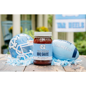 A jar of Cackalacky - Carolina Tar Heels BBQ Sauce is placed on a wooden surface, alongside a light blue football helmet and football. The display is artistically enhanced with scattered blue and white paper shreds, highlighting the vibrant appeal of this savory tomato-based barbecue sauce from Cackalacky.