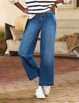 A person wearing a striped sweater, lightweight FRANK AND EILEEN - WESTPORT ITALIAN WIDE LEG TROUSER, and white sneakers stands on a brick path, holding a straw hat. The casual scene is enhanced by the textured backdrop, offering a relaxed yet stylish vibe.