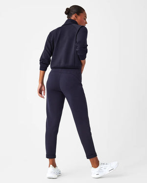 A person stands against a white background, smiling and looking down with one hand touching their hair. They are wearing a matching light beige SPANX AIRESSENTIALS TAPERED PANT and sweatshirt set made from ultra drapey, lightweight spacer fabric, paired with white sneakers.