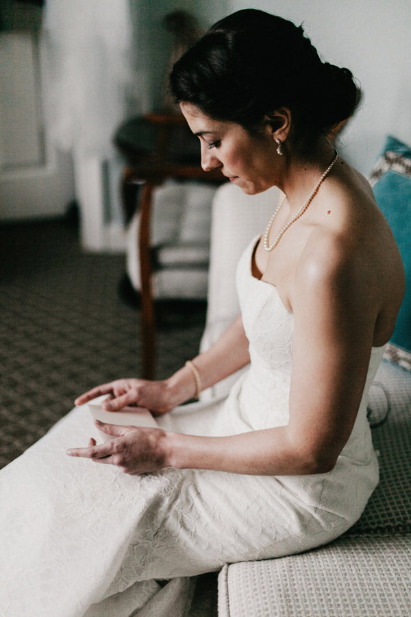 A bride in a strapless white wedding dress sits on a patterned couch, looking down at her hands. She wears a pearl necklace and bracelet, with her dark hair pulled back in an elegant updo.