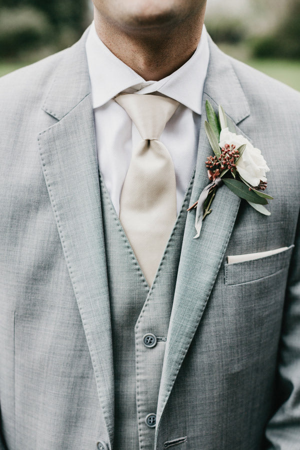 A man wearing a light gray suit, cream-colored tie, and white dress shirt with a white rose boutonniere pinned to his lapel. His face is partially out of frame.