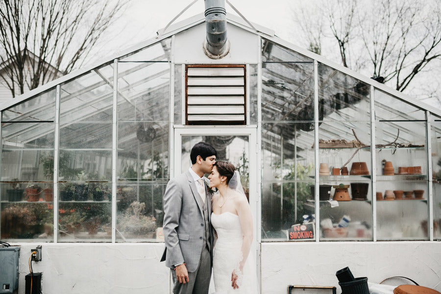 A bride and groom stand close together, touching foreheads, in front of a glass greenhouse filled with plants and terracotta pots. The groom wears a light gray suit; the bride wears a strapless white dress.