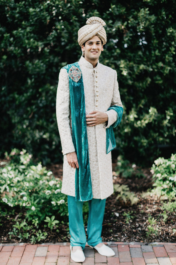 A man in a cream-colored traditional sherwani with ornate buttons, teal pants, and a matching teal stole stands outdoors on a brick path, wearing a cream turban, with greenery in the background.