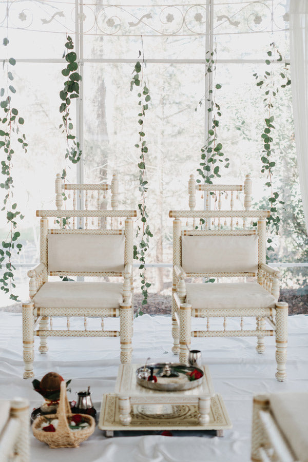 Two ornate ivory chairs with gold accents are placed side by side on a white floor, with greenery hanging in the background and ceremonial items arranged on trays in front of them.