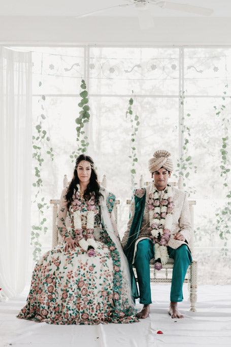 A bride and groom in traditional South Asian wedding attire sit side by side on ornate chairs, adorned with flower garlands, against a backdrop of greenery and sheer curtains.
