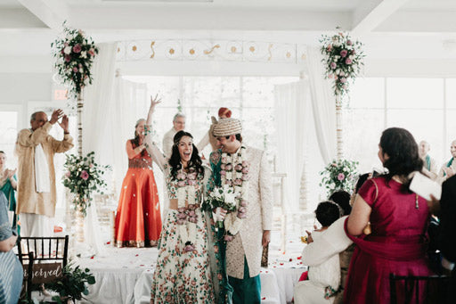 A joyful couple in traditional Indian wedding attire walk down the aisle, smiling and celebrating, surrounded by applauding guests and floral decorations in a bright, elegant venue.