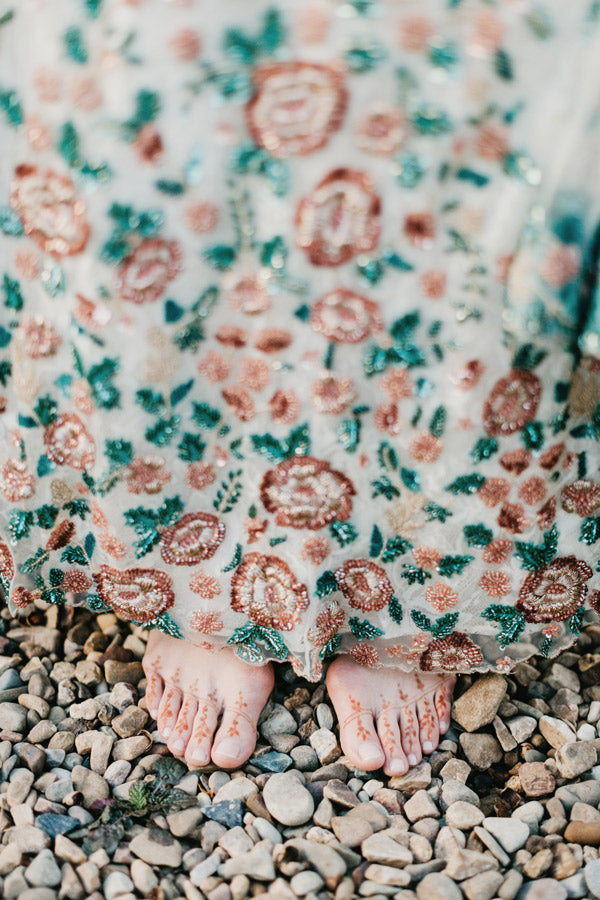 Bare feet with henna designs peek out from under an embroidered, floral-patterned dress, standing on light-colored pebbles.