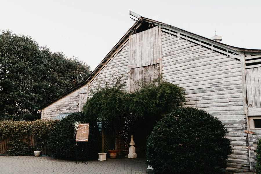 A rustic wooden barn with weathered siding, surrounded by green bushes and trees. The entrance is adorned with climbing plants and string lights, and a decorative sign stands near the doorway on a paved path.
