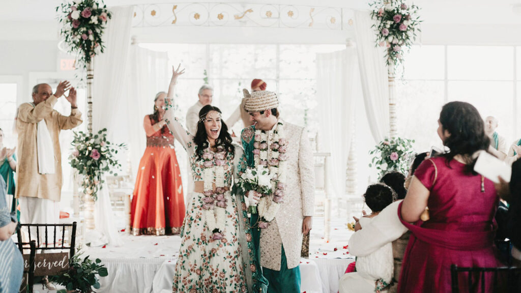 A joyful bride and groom in traditional Indian attire walk down the aisle, smiling and holding hands, surrounded by cheering guests and floral decorations on a bright, festive wedding stage.