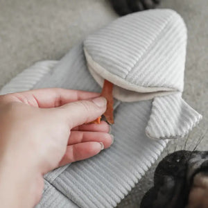 A person's hand places a small treat inside a soft LAMBWOLF COLLECTIVE - TROUT DOG TOY, shaped like a shark, which is laying on a carpet. Nearby, a dog's paw and part of its nose are visible, suggesting the dog eagerly waiting to engage with this enrichment play object from LAMBWOLF COLLECTIVE.