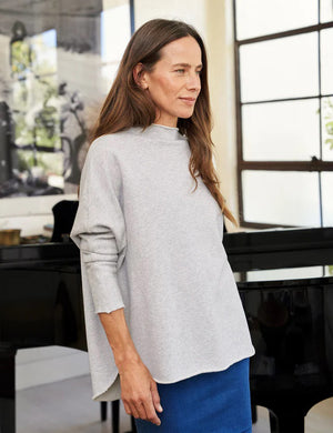 A woman with long brown hair stands smiling near a black grand piano in a modern, well-lit room. She wears the FRANK AND EILEEN - EFFIE LONG SLEEVE FUNNEL NECK CAPELET from FRANK & EILEEN paired with blue jeans. A large window and a lamp are in the background, enhancing the cozy winter warmth of her stylish ensemble.