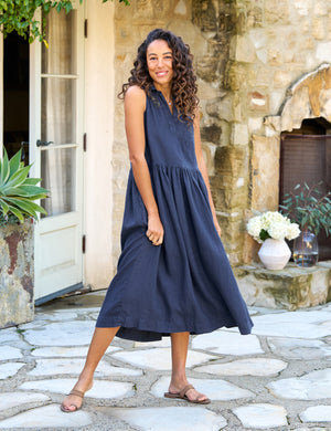 The FRANK & EILEEN - Daphne Sleeveless Popover Dress is a sleeveless, dark blue midi dress with a fitted bodice, collared neckline, and a lightly pleated, flowing skirt. Crafted from washed linen, the dress also features handy side pockets. Displayed on a white background.